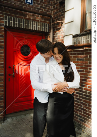 Young loving couple embracing in front of a vibrant red door, showcasing affection and joy 130110555