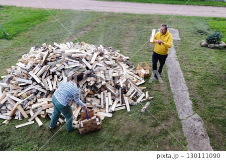 Middle-aged husband and wife pick up firewood from large pile and stack it into wicker baskets, view from above. Middle-aged husband and wife pick up firewood from large pile and stack it into wicker baskets, view from above. 130111090