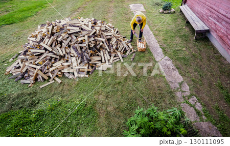 Gray haired man takes firewood from pile near country house and puts it into large wicker basket. 130111095