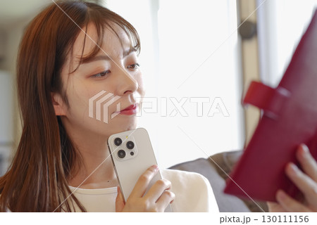 ソファに座って手帳の予定を見る女性 130111156