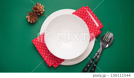 Christmas table setting with empty plate, golden pine cones, and red napkin on green background 130111544