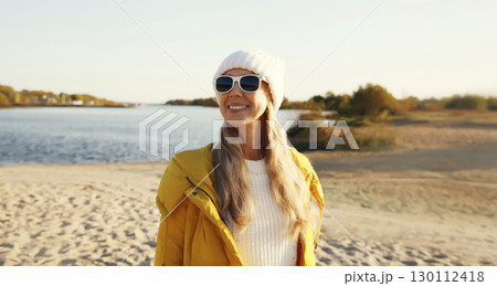 Happy woman standing on sunny beach looking away, wear jacket, sea coast 130112418