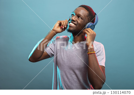 Cheerful male individual looking up and enjoying his favorite songs against a blue background. Happy african american man in casual attire, touching his wireless headphones and listening to music. Cheerful male individual looking up and enjoying his favorite songs against a blue background. Happy african american man in casual attire, touching his wireless headphones and listening to music. 130114070