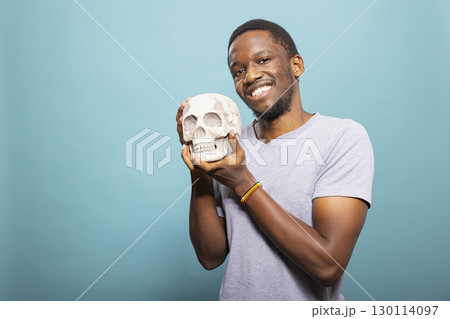 Cheerful black male person stands and grasps a skull close to him, posing for the camera in a studio. Portrait of a happy african american man holding a human skeletal head. 130114097