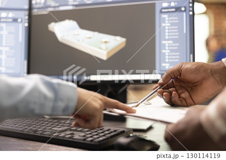Selective focus on multiethnic developers with a clipboard and pen at desk, discussing prototypes. They are collaborating on design adjustments with CAD software displayed on a desktop pc monitor. 130114119