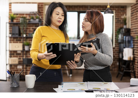 Two female colleagues standing and sharing their strategy ideas in brick wall office. Businesswoman holding a clipboard, comparing project notes with her coworker using a tablet in modern workspace. Two female colleagues standing and sharing their strategy ideas in brick wall office. Businesswoman holding a clipboard, comparing project notes with her coworker using a tablet in modern workspace. 130114123
