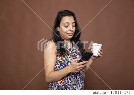 Female individual with disposable cup, using her smartphone, chatting and browsing on social media. Indian woman holding disposable cup and mobile device as she stands against isolated background. 130114173