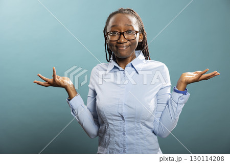 Confused black woman looking at camera with pursed lips and shrugged shoulders. African American female individual spreading her hands, expressing uncertainty with I dont know gesture. Confused black woman looking at camera with pursed lips and shrugged shoulders. African American female individual spreading her hands, expressing uncertainty with I dont know gesture. 130114208