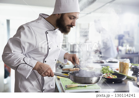 Steam rises from a sizzling pan as a male head chef prepares a tasty recipe in a modern restaurant kitchen, working at the counter on a gourmet dish. Passion for culinary arts. 130114241