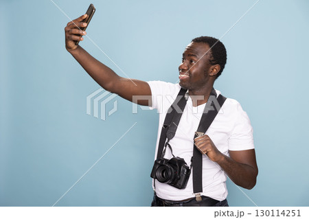 Young black man taking a selfie with his cellphone. African american male individual, with a camera around his neck, grips his suspenders strap while recording a video on his mobile device. 130114251