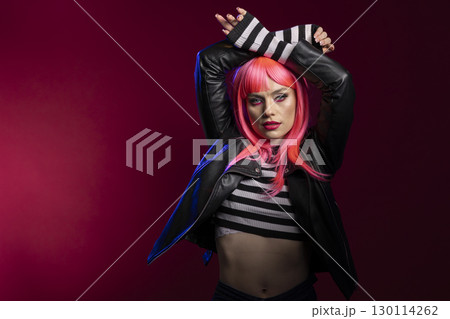 Fashion forward woman with vibrant pink hair and striped crop top poses in dramatic scene. Stylish female model with arms above head, confidently stands in artistic studio lighting. Fashion forward woman with vibrant pink hair and striped crop top poses in dramatic scene. Stylish female model with arms above head, confidently stands in artistic studio lighting. 130114262