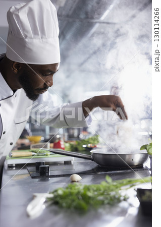 African american head chef sprinkles rosemary over a gourmet dish slow cooked on the stove, balanced gastronomy with fresh herbs. Kitchen counter filled with raw materials and fresh ingredients. 130114266