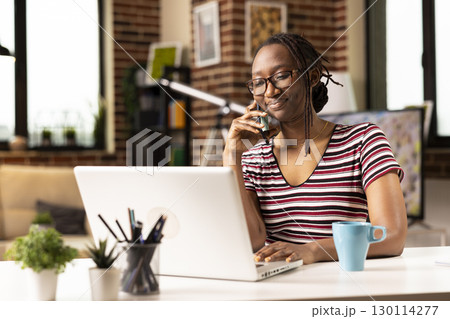 Self employed woman sits at desk, multitasking, speaks with clients on phone and updates data on laptop. Smiling female freelancer uses mobile device while working from home on personal computer. Self employed woman sits at desk, multitasking, speaks with clients on phone and updates data on laptop. Smiling female freelancer uses mobile device while working from home on personal computer. 130114277