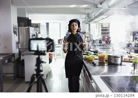 Professional chef in uniform recording a cooking tutorial in a modern kitchen, sharing expert techniques and recipes for an online audience. Woman filming every step of food prep. 130114284