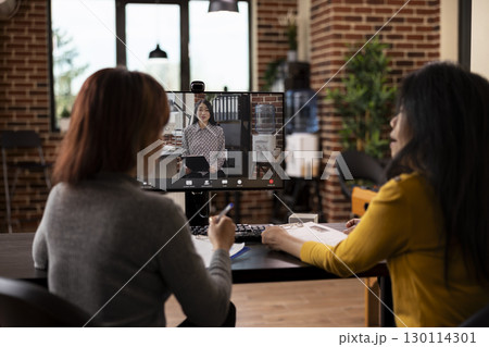 Female colleagues seated at desk, on a video conference call with their asian manager seen on desktop monitor. Businesswomen in office using computer for a virtual meeting with potential partner. Female colleagues seated at desk, on a video conference call with their asian manager seen on desktop monitor. Businesswomen in office using computer for a virtual meeting with potential partner. 130114301
