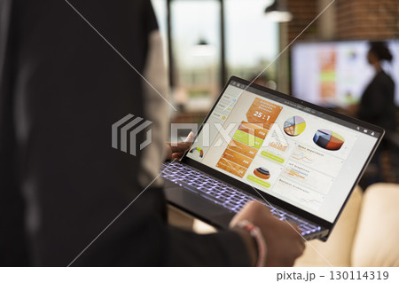 Selective focus on device screen displaying business analytics charts. Rearview closeup of african american person walking in office, reviewing financial graphs and evaluating kpi on laptop. Selective focus on device screen displaying business analytics charts. Rearview closeup of african american person walking in office, reviewing financial graphs and evaluating kpi on laptop. 130114319
