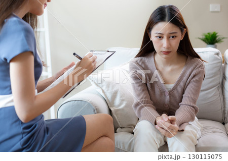 Asian women looks anxious and silent while sitting on a sofa during a mental health consultation as female therapist takes notes, showing emotional struggle. Therapy session concept. Asian women looks anxious and silent while sitting on a sofa during a mental health consultation as female therapist takes notes, showing emotional struggle. Therapy session concept. 130115075