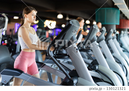 Young woman stands on simulator, performs fitness task using an elliptical simulator Young woman stands on simulator, performs fitness task using an elliptical simulator 130121321