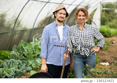 Man and woman with rakes in hands inside greenhouse with vegetable harvest 130121421