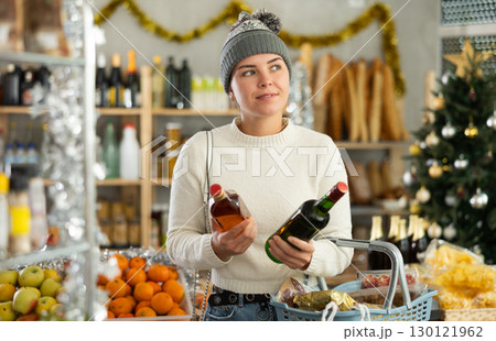 Positive woman choosing whiskey for Christmas feast in decorated store 130121962