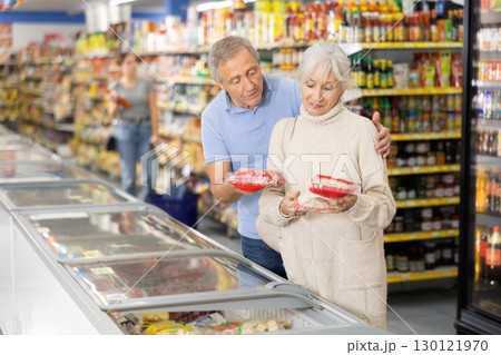 Aged couple looking at goods in refrigerated display case in food store Aged couple looking at goods in refrigerated display case in food store 130121970