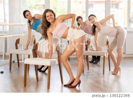 Leaning on chair seat with hand foot, young woman performs lap dance during group workout. Leaning on chair seat with hand foot, young woman performs lap dance during group workout. 130122169