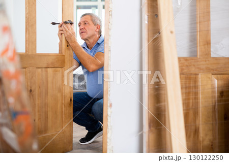 Professional middle-aged repairman installing a door handle on a door during renovation, indoors Professional middle-aged repairman installing a door handle on a door during renovation, indoors 130122250
