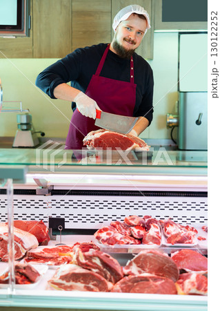 Young bearded butcher slicing fresh beef for customer at counter 130122252