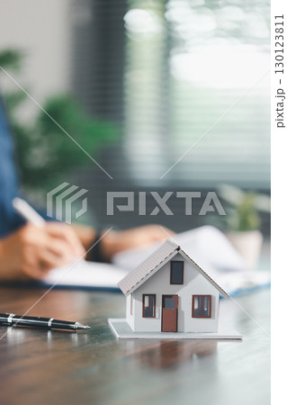 Real estate agent and customer are discussing contract to buy house, with house model in background symbolizing real estate, insurance, and loan arrangements involved in process. 130123811