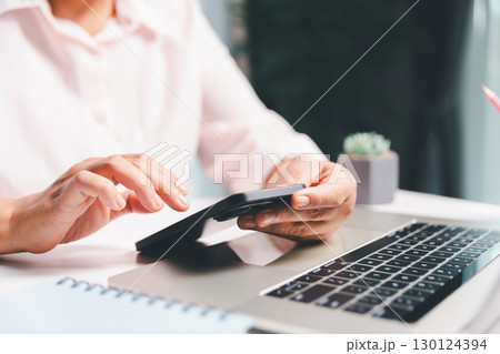 Close-up shot, woman sits at a wooden table using her smartphone, with her finger touching the phone screen as she navigates mobile banking, online shopping, and social media networks. 130124394