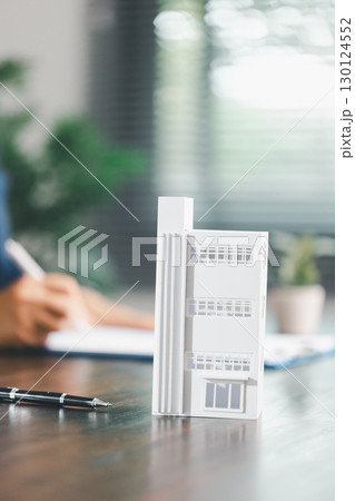 Real estate agent and customer are discussing contract to buy house, with house model in background symbolizing real estate, insurance, and loan arrangements involved in process. 130124552