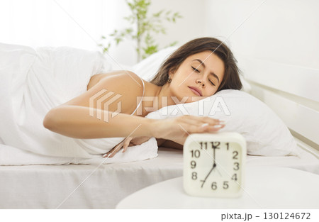 Peaceful young woman sleeping comfortably in bed with alarm clock nearby, resting early in morning. 130124672