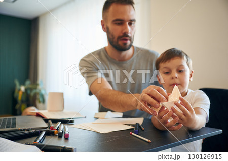 Playing with origami boat. Father and son are at home by the table Playing with origami boat. Father and son are at home by the table 130126915