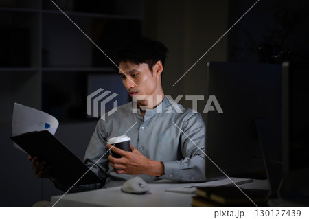 Businessman working late at night holding coffee cup and reading documents at his desk 130127439