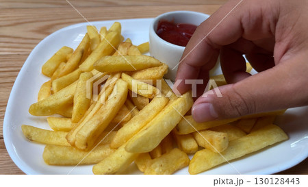 A child's hand takes pieces of french fries from a plate and dips them in tomato ketchup 130128443