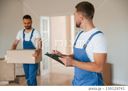Writing in notepad, holding box. Two moving service employees in a room 130129741