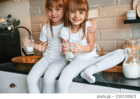 Embracing each other. Happy two little girls are drinking milk while sitting on the table on the kitchen Embracing each other. Happy two little girls are drinking milk while sitting on the table on the kitchen 130130021