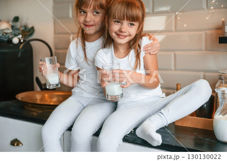Embracing each other. Happy two little girls are drinking milk while sitting on the table on the kitchen Embracing each other. Happy two little girls are drinking milk while sitting on the table on the kitchen 130130022