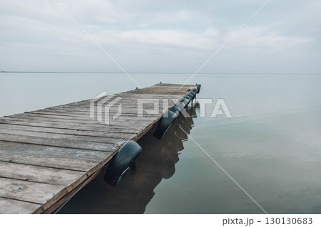 wooden pier stretching on calm still lake under soft cloudy sky, peaceful waterscape scene with muted colors and quiet atmosphere, travel inspiration, nature therapy, wellness retreats 130130683