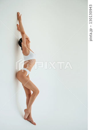 Leaning on the wall, with hands up. Woman in white underwear is against white background 130130943