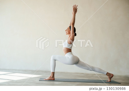 Doing warrior yoga asana exercise. Fitness woman is indoors in white clothes 130130986