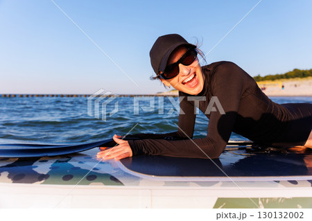 Smiling woman enjoys surfing on Baltic Sea waves, laughing joyfully lying on board with splashing water around. Bright sunlight and her laughter reflect pure joy, freedom and vibrant summer adventure 130132002