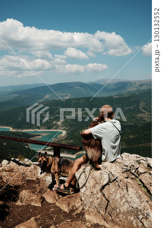 Man sitting on rock with two dogs German and Australian Shepherd overlooking stunning mountain valley and turquoise lake in Stara Planina, Serbia. Travel and hiking with pets concept 130132552