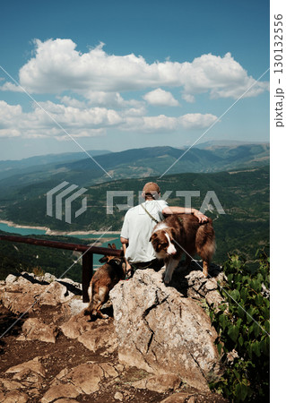 Man sitting on rock with two dogs German and Australian Shepherd overlooking stunning mountain valley and turquoise lake in Stara Planina, Serbia. Travel and hiking with pets concept 130132556