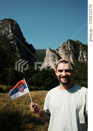 Man smiling while holding a Serbian flag in front of tall rocky mountains and blue sky. Stara planina national park Man smiling while holding a Serbian flag in front of tall rocky mountains and blue sky. Stara planina national park 130132570