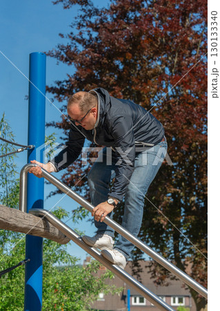 Man in jeans and jacket climbs a modern playground setup. Sunny weather, outdoor fitness, and a relaxed lifestyle moment for adult recreation in urban surroundings. Man in jeans and jacket climbs a modern playground setup. Sunny weather, outdoor fitness, and a relaxed lifestyle moment for adult recreation in urban surroundings. 130133340