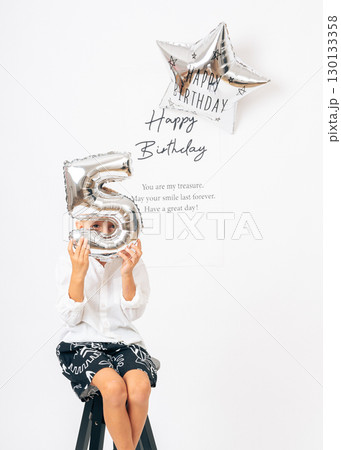Cheerful five-year-old boy in white shirt posing with silver balloon on birthday. Ideal for childrens events, birthday cards, family content, and editorial or advertising use 130133358
