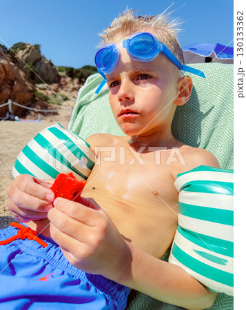 Young child relaxes on a beach with inflatable arm bands, wearing blue goggles and holding a piece of watermelon. Sunlight highlights the scene, capturing a moment of summer leisure. 130133362