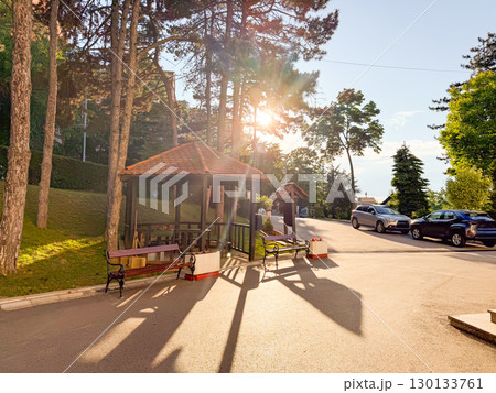 Evening sun shining through pine trees and wooden gazebo with benches near the roadside. Relaxation, summer atmosphere, peaceful rest and connection with nature. 130133761