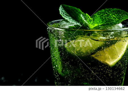 A vibrant cocktail filled with ice, mint leaves, and lime is elegantly displayed against a smooth black backdrop, highlighting its refreshing appeal 130134662